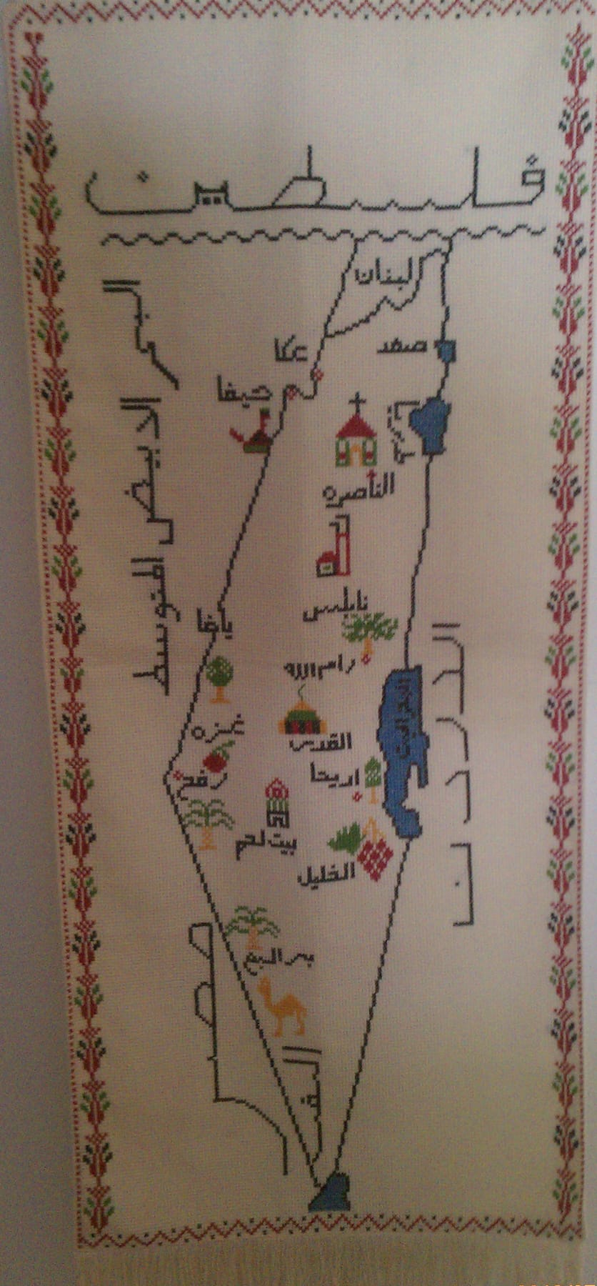 Embroidered tatreez map of Palestine: cross-stitch on white cloth with a red, black and green floral border, the word فلسطين stitched in black above the map, coastal towns from Acre to Gaza marked with small stitched landmarks — a church, a mosque, olive and palm trees, a camel — and Lake Tiberias and the Dead Sea in blue.